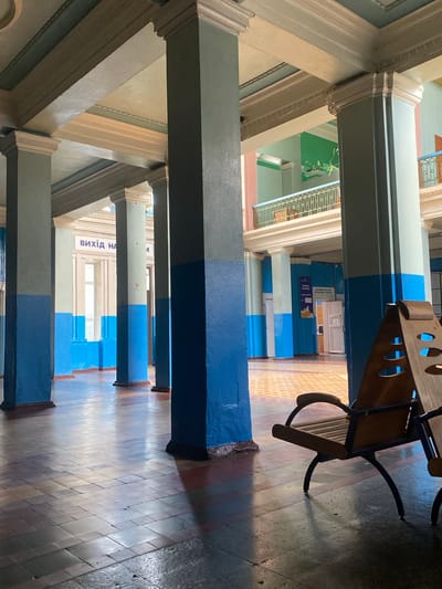 The image shows a long, open hallway with a series of tall, painted columns. The columns are painted in two horizontal sections: the lower section is a bright blue, and the upper section is a muted greenish-gray. The ceiling is ornate with architectural details and painted in pale green and cream colors. Sunlight streams in from large windows on the left side of the hallway, casting long shadows across the tiled floor. The floor is made of large, square tiles in various shades of brown and gray, with a patterned inlay in the distance.

In the foreground on the right, a wooden bench with a dark metal frame is visible. It has a slatted seat and backrest, and its legs are curved and thin. Another bench is partially visible to the right of the first.

Further down the hallway, on the left wall, there is signage with Ukrainian text, including the words "ВИХІД НА" (EXIT TO). Beyond the columns, the hallway opens up to a second level with a railing. On the right side of the hallway, there are doors and more signage with Ukrainian text, including "ГРАДНО" and what appears to be a logo.

The overall impression is of an old, possibly institutional building, like a train station or a public institution, located in the town of Romodan, Ukraine. The building appears to be well-maintained but dated in its style and decor. There are no people visible in the image.