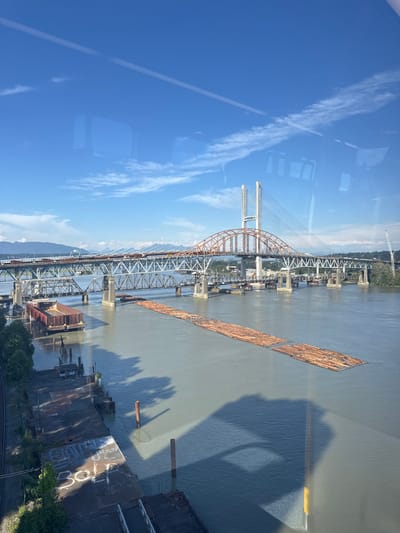 The image depicts a wide, calm river with multiple bridges spanning across it, a common sight in New Westminster, Canada. The foreground features a concrete pier with graffiti that reads "BOMB" and "BOLD." Further out on the river are two barges loaded with lumber, presumably awaiting transport. The middle ground is dominated by two large bridges: a lower, older truss bridge and a higher, newer cable-stayed bridge with distinctive white towers. The background shows a tree-lined shore and distant mountains under a bright blue sky with scattered white clouds. Reflections of windows and possibly clouds are visible on the surface of the water and the sky, suggesting the photo was taken from an elevated interior perspective.