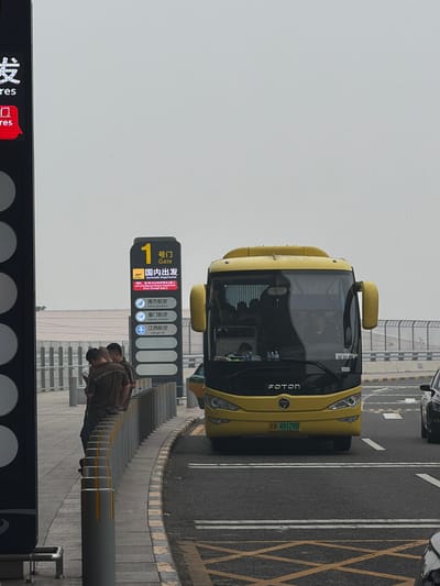 The image depicts an outdoor scene, likely at an airport, given the signage and the presence of a bus. The weather appears overcast. In the foreground, on the left side, a tall black sign with illuminated white characters is visible, with "ures" being the only decipherable English text. A red rectangular sign with Chinese characters and the word "res" is below it. Further to the left, several vertical circular elements are visible on the black sign.

To the right of the black sign, a vertical illuminated signpost is prominently displayed. It has a yellow rectangular section at the top with the number "1" and Chinese characters, followed by "Gate". Below this, it reads "国内出发" (Domestic Departures) in large characters, and then in smaller text, "Int'l/HK/Macao/Taiwan Departures Enter through Gate 3". Underneath this section, logos and names for "China Southern", "Xiamen Airlines", and "Jiangxi Air" are displayed vertically.

In front of the signpost, three men are standing and looking down at a phone. Two of them are wearing similar brown shirts. They are positioned near a row of cylindrical bollards with yellow tops.

A yellow bus, with "FOTON" branding on the front, is parked on the right side of the image, facing towards the viewer. The bus has its headlights on. The license plate reads "A 49128D". Inside the bus, passengers can be seen, and the driver is in the driver's seat. Another vehicle, a dark-colored car, is partially visible to the right of the bus, also on the road.

The ground consists of asphalt roads and a paved pedestrian area with yellow and white lane markings. A metal railing runs along the pedestrian walkway. The background shows a blurred view of buildings and a fence.

The scene appears to be located in Guangyang District, China, as indicated by the Chinese signage and the nature of the airport-related infrastructure.