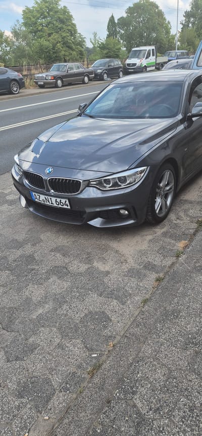 This image shows a parked dark gray BMW in an urban setting, likely in Mainz, Germany. The car is positioned at an angle, with its front right side facing the viewer. It is parked on a sidewalk paved with hexagonal concrete tiles. Beyond the sidewalk, a road is visible with white lines. In the background, several other vehicles are parked in a line, including a white van with green and red accents and an older model dark sedan. Trees and foliage line the area behind the parked cars, suggesting a roadside location. The lighting indicates it is daytime, possibly late afternoon, with scattered clouds visible in the reflection on the car's hood. The license plate of the BMW is partially visible, reading "AZ NI 664". There are no people visible in the image. The overall scene depicts a quiet moment of parked vehicles on a typical urban street.