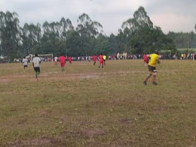 A soccer game unfolds on a muddy field under a cloudy sky. Players in red and yellow jerseys compete, while others in white watch nearby. A simple goalpost stands at one end. Spectators line the edges of the field, creating a visible boundary between the game and the surrounding trees. The focus is on the players and their activity on the field.