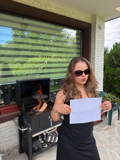 In Priseltsi, Bulgaria, a young woman with brown, wavy hair, wearing sunglasses and a black dress, stands in front of a black barbecue grill. The grill contains burning logs and is positioned on a patio with light-colored stone tiles. Behind the woman and grill is a house with a textured white stone facade and a large window covered by green and white blinds. The woman holds a white piece of paper with "Collective MEMORY" and an infinity symbol drawn on it in blue ink. The scene is outdoors during the daytime, with trees and greenery visible in the background and a cloudy sky.