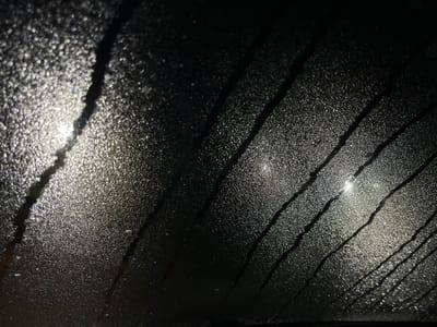 The image displays a close-up, abstract view of a surface covered in numerous tiny water droplets, likely condensation or heavy moisture on a pane of glass such as a window or windshield. The overall scene is dark, suggesting nighttime or a low-light environment.
Bright, diffused light sources are visible through or reflecting off the water-laden surface. These lights appear as soft, glowing areas of white and light grey, scattered and blurred by the moisture, creating a bokeh effect. The individual water droplets act as tiny reflective points, giving the surface a shimmering, granular texture wherever the light catches them.
Running diagonally across the textured, wet surface are several distinct, darker streaks. These streaks are noticeably clearer than the surrounding area, indicating paths where water has either coalesced and flowed, or where the surface has been wiped, clearing the condensation. These lines vary in thickness and appear somewhat parallel, extending from the top left to the bottom right of the frame, and from top right towards the bottom left in some areas. They stand in stark contrast to the illuminated, droplet-covered regions.
The dominant colors are shades of black and dark grey in the unlit areas and within the streaks, contrasting with the bright white and light grey of the illuminated water droplets. There is no discernible text or specific objects visible beyond the pattern of light, moisture, and streaks on the surface. The image evokes a sense of looking out from a confined space into a dark, wet environment with distant lights.