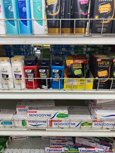 A shelf in a store in Kecamatan Piyungan, Indonesia (latitude: -7.837, longitude: 110.474) with various toiletries and personal care products. On the top shelf are several bottles of shampoo, including "Pantene" and "Rambut Rontok" brands. The middle shelf contains several bottles of hair care products, including "Men's Biore", "Natura", and "Cool Energy" brands. The bottom shelf is stocked with several tubes of toothpaste, including "Sensodyne" and "Ciptadent" brands. The shelves are made of metal with a white background. The lighting is fluorescent. The products are arranged in a neat and orderly manner. The shelves are relatively empty. There are no people in the image.