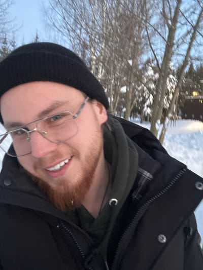A fair-skinned man with red hair and a trimmed beard smiles at the camera. He is wearing glasses and a black beanie. His attire includes a dark green hoodie under a black winter coat. The background shows a snowy outdoor scene with bare tree branches and a building in the distance. The image is rotated, presenting the subject at an angle.
