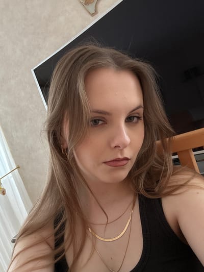 A close-up selfie captures a young woman with fair skin, light brown hair, and a neutral expression. She has green eyes and appears to be wearing subtle makeup, including eyeliner and lipstick. A black tank top is visible, adorned with two gold necklaces. The background includes a black surface, a wooden chair, and a light-colored wall with a textured pattern. There is no indication of the location being in Truchtersheim, France.