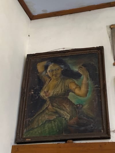 This image depicts an indoor corner located in Mojokerto, Indonesia. A framed oil painting is prominently displayed on a white wall. The painting portrays a woman with dark hair and a medium complexion, depicted from the waist up, wearing a light-colored, low-cut garment that exposes cleavage. Her left arm is raised, with her hand positioned near her head, while her right arm is bent, adorned with a bangle, and situated above a small, dark, lamp-like object. The painting's background is dark, featuring a central greenish-blue glow. The canvas itself shows significant cracking and flaking of paint, particularly on the left side. The artwork is enclosed within a dark, ornate wooden frame that exhibits signs of surface wear and minor chipping. A thin wire for hanging is visible above the frame. The white wall transitions to a light blue ceiling or upper wall section, separated by a dark wooden trim. This ceiling area displays multiple water stains and discoloration. A segment of a wooden shelf or beam is visible immediately below the painting. No individuals are observed interacting within the depicted environment.
