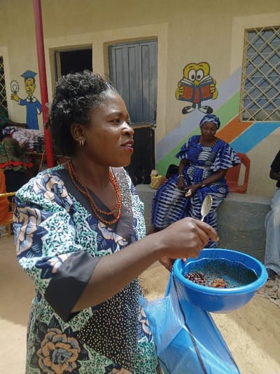 This image depicts an outdoor scene in Masvingo, Zimbabwe. In the foreground, a woman with dark skin, short, dark, coily hair, gold hoop earrings, and a multi-strand orange beaded necklace wears a patterned dress featuring teal, light blue, black, white, and orange floral and abstract designs with black sleeve cuffs. Her left arm is extended, holding a silver spoon above a blue plastic bowl. The bowl contains dark granular material, small reddish-brown berries, and a brownish lump, and is situated within a translucent blue plastic bag. Her mouth is slightly open, and she looks towards the right.

In the mid-ground, a second woman, also with dark skin, wearing a blue and white patterned head covering and a matching dress, is seated on a red plastic chair, observing the foreground figure. A third individual is partially visible on the far right.

The background consists of a light-colored building wall adorned with two murals: one depicting a figure in a blue graduation gown holding a lightbulb on the left, and another showing a yellow cartoon character reading an open book on the right. Below the reading character, the wall features diagonal stripes of orange, green, and light blue. A dark gray two-panel door is partially open on the wall, and a window with a metal security grill is visible to the right of the reading mural. The ground is light-colored dirt or sand, and a concrete step or platform is present where the seated woman is positioned. A red vertical pole is partially visible on the far left of the frame.