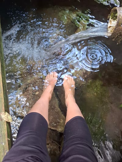 The image shows a pair of bare feet and lower legs dipped in water, likely a natural spring or small stream. The person is wearing black leggings that are cropped below the knee. A pipe drains water into the pool of water, creating ripples and bubbles. The water is clear enough to see the bottom, which appears to be made up of rocks and pebbles. The scene is located in Khlevnoye, Russia.