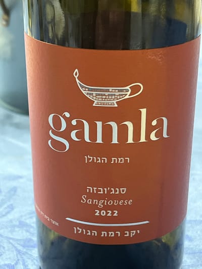 A wine bottle label, predominantly brown, features the name "Gamla" in a large, elegant font. Below the name, in Hebrew, reads "Ramat Hagolan." To the right, "Sangiovese" and the year "2022" are printed. At the top right corner, also in Hebrew, it says "Yekev Ramat Hagolan". A stylized drawing of an ancient oil lamp adorns the label to the left of the name. The bottle itself appears to be green glass.