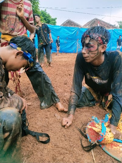 The scene takes place in an open area covered in dirt. There is a blue tarp behind the individuals in the foreground. The area appears to be a performance area, with a young male sitting on the ground with his right leg extended and left leg bent. He is wearing black pants and a black shirt with a white logo and is staring off to the side. His face is painted with black and red markings. His hands are touching the ground, and a yellow, blue, and red drum is lying to his right. He is likely performing a traditional ritual dance. There is a second young male sitting in the foreground with his back to the camera. His legs are extended and his arms are resting on his knees. He is wearing a black and white shirt and black pants. There are two other figures in the background: a young male who appears to be watching the performance and a second young male who is standing to the right of the scene. The location is in Kecamatan Kedawung, Indonesia, with the coordinates -7.494, 111.07.  The image shows a group of individuals during a traditional performance.