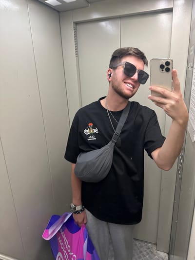 In the city of Mirny, Russia, a young man is taking a selfie in an elevator. He is wearing a black t-shirt with a cartoon graphic, gray sweatpants, sunglasses, and wireless earbuds. He has a silver necklace and a watch on his left wrist, and a gray crossbody bag slung over his shoulder. In his left hand, he holds a brightly colored shopping bag with text on it. The elevator walls are a light gray metallic material, and the floor is textured metal.