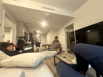 The image depicts an indoor scene within a modern apartment in Washington, United States, specifically a combined living room and kitchen area. The perspective is low, from the viewpoint of a person seated on a sofa.
Five individuals are present. In the immediate foreground, the lower torso and legs of a person are visible on the right, clad in dark blue athletic pants and light-colored shoes, with their feet resting on a coffee table. To the left, a young male with dark curly hair and a mustache is reclining on a light-colored sofa, wearing a dark long-sleeved shirt, with his left hand near his head. His legs, covered by light-colored pants, are propped on a cushion.
In the mid-ground, a young male with dreadlocks or long hair is standing in the kitchen area, facing left but looking slightly towards the viewer. He wears a grey t-shirt and dark shorts, utilizing crutches under both arms, and has a brace or bandage on his right knee. To his right, another young male with dark hair is seated at a light-colored dining table, wearing a light brown long-sleeved shirt and dark pants. In the background, partially visible through a doorway on the right, a fifth young male with dark hair stands, wearing a light brown jacket or shirt and light pants, observing the scene.
The environment features light-colored walls, a light-colored wooden or laminate floor, and overhead artificial lighting from ceiling fixtures and recessed lights. The kitchen area includes light-colored upper cabinets, dark lower cabinets, a dark countertop, and stainless steel appliances including a microwave. A large, dark flat-screen television is mounted on the wall to the right. A rectangular wooden coffee table with a striped pattern holds small items including a circular coaster, a green object, and a red object. No direct interactions or specific activities beyond presence in the shared space are discernible.