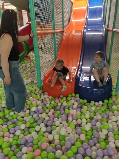 This is a media file showing a ball pit and slides in a children's play area. Three individuals are visible. Two young boys are on the slides. One boy is at the top of a blue slide, and another boy is halfway down an orange slide. A woman is partially visible in the foreground, appearing to reach up towards the structure. The ball pit is filled with a mixture of green, white, and light purple balls. The scene appears to be indoors, likely in a play center or arcade. The lighting suggests it is daytime. There are no visible text elements in the image. The overall atmosphere seems playful and energetic.