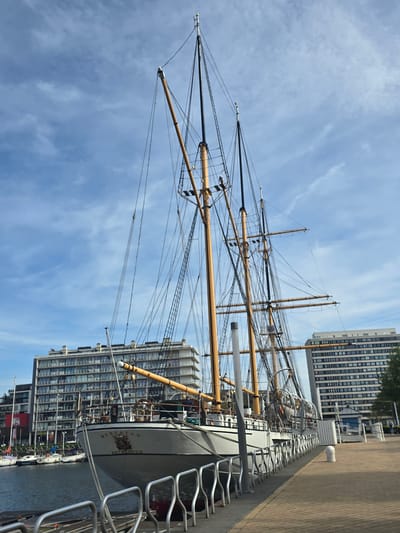 The image shows the Mercator, a three-masted barquentine, docked at Ostend, Belgium. The ship is seen from the side, its masts and rigging extending high against a blue sky with light clouds. A modern building is visible in the background, next to the docked ship. A metal railing runs along the right side of the frame, suggesting a walkway or pier. The ship is docked beside water.