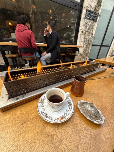 The image depicts an outdoor cafe setting from a first-person perspective, with a wooden table in the foreground. Occupying the middle ground is a rectangular, dark metal fire pit with numerous circular perforations along its sides, emitting multiple bright orange flames. To the right of the fire pit, on the wooden table, sits a white ceramic coffee cup with a multicolored floral pattern on both the cup and its matching saucer, containing a dark liquid and residual sediment. Behind the cup is a small, unlidded, hammered copper pot. Further to the right, a silver-colored, ornate metal lid with a finial rests on the table.
In the background, beyond the fire pit, two adult males are seated opposite each other at a high-top table beside a large glass window facade. The man on the left, viewed from the back, wears a red hooded sweatshirt and blue jeans. The man on the right, seen in profile, wears a dark long coat over a light shirt and light grey trousers; he is actively smoking a cigarette, holding it to his mouth with his right hand. Reflections of interior lighting and elements are visible in the glass. Adjacent to the window facade on the right is a light-colored stone wall partially covered by climbing green vines. Affixed to this stone wall is a blue street sign, partially obscured, clearly displaying "شارع الحميدية | HAMIDIYEH ST." in both Arabic and English, indicating the location in Amman, Jordan. The overall lighting is subdued, suggesting an overcast day or late afternoon.