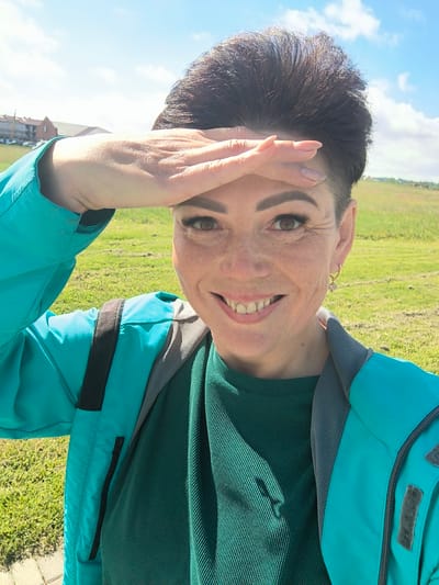 A woman stands in a grassy field under a bright, cloudy sky, possibly in Laskino, Russia. She is taking a selfie and shielding her eyes from the sun with her left hand. The woman has short, dark hair, freckles, and is wearing a teal jacket over a dark green ribbed shirt. She is smiling and has visible makeup, including mascara and lipstick. A small, geometric earring is visible on her right ear. The background shows a green, rolling hill with some buildings in the distance. The lighting suggests it is daytime, and the presence of a jacket implies it is not extremely hot weather.