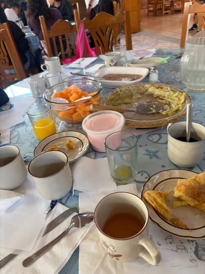 The image shows a breakfast table setting in what appears to be a private home. The location may be Santa María Zacatepec, Mexico. Multiple people are seated around a table, partially visible in the background. The tabletop features a blue and white patterned tablecloth. Various food items are present, including a bowl of scrambled eggs, a plate with pastries, a bowl of cut melon, and dishes containing crumbs. There are also various beverages: orange juice in a glass, a pink smoothie, and a mug with coffee. Utensils, including spoons and knives, are laid out on napkins next to the place settings. There are at least three mugs and several glasses on the table. The room is lit with natural light coming from an unseen window.