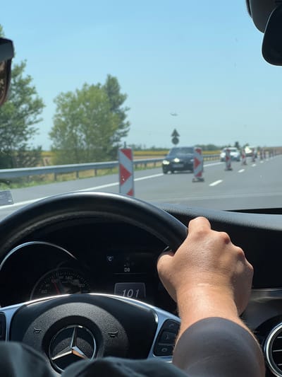 The image shows a first-person perspective of someone driving a car. The driver's hand is on the steering wheel, which has a Mercedes-Benz logo. The dashboard is visible, displaying a speed of 101 km/h and other indicators like "R", "N", "P", and "D8". The road ahead is visible through the windshield, with traffic cones and construction barriers lining the right side of the road, suggesting ongoing road work. Trees and a bright blue sky are also visible. The lighting suggests it is daytime, likely late morning or afternoon, with clear weather. The scene appears to be a highway or a major road in a rural or semi-rural area, given the presence of trees and open sky. The overall impression is one of normal driving activity on a sunny day with some road construction ahead.