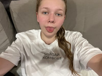A young woman is lying down on a sofa in a room, looking up at the camera. She has light brown hair that falls around her face and shoulders. She is wearing a white t-shirt with black text printed on it. The text is in Russian and reads "СОЗДАЙ КОМФОРТ", which translates to "Create Comfort". Her facial expression is playful as she sticks her tongue out slightly. The lighting in the room appears to be artificial, suggesting it might be evening or indoors during the day. The background consists of a textured sofa and pillows, common in a domestic setting. The overall mood of the image is casual and lighthearted, captured in a selfie-style shot. The provided location context places this scene in Klepiki, Russia.