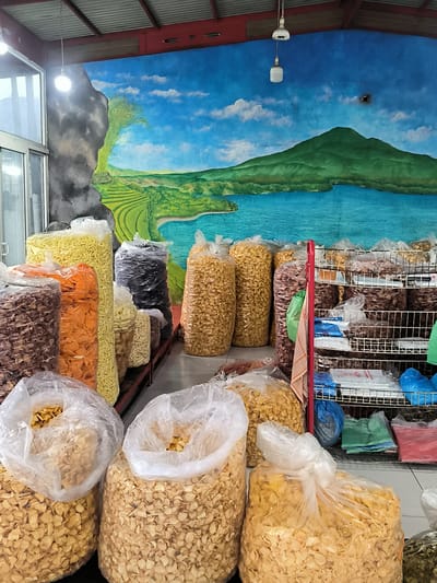 This image captures the interior of a snack shop or market stall, likely located in Sukamulia, Indonesia. The dominant background feature is a vibrant mural painted across the back wall, depicting a picturesque mountainous landscape. The mural showcases a large green mountain, a sprawling blue lake, and verdant terraced rice fields, all under a bright blue sky dotted with white clouds. A dark, rocky formation is painted on the left side of the mural.

The foreground and midground are filled with a large assortment of snacks, predominantly packed in clear plastic bags. These bags, varying in size, contain different types of crackers or chips, distinguishable by their shapes, colors (including yellow noodles, orange slices, dark brown and black varieties, and light beige shapes), and textures. Many bags are arranged standing upright on the floor or on low platforms, while others are piled.

On the right, a red metal shelving unit with wire racks displays more snack bags and various folded plastic bags in blue, green, and red, along with some white packages. A red and white checkered cloth is draped over the side of this shelving unit. The ceiling features exposed red and grey metal beams with corrugated roofing, and bare lightbulbs provide artificial illumination. A glass door or window is visible on the far left.

No people are present in the scene, which focuses entirely on the display of goods for sale. The snacks appear to be traditional Indonesian *kerupuk* or *kripik*. There is no visible text on any of the products or packaging.