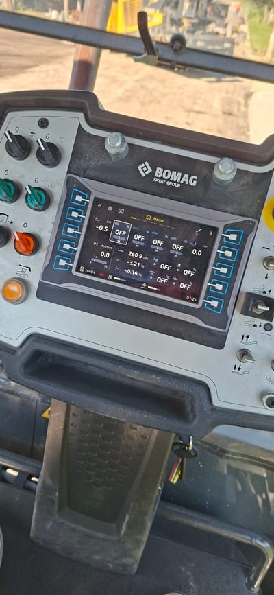 An interior shot of a Bomag Fayat Group compactor's control panel is captured from a slightly elevated, forward-facing perspective. The panel features a digital display screen showing various operational parameters, including "OFF" for multiple functions, speed readings like "0.0 m/min," and measurements in "cm" and "%." Several physical control knobs and buttons are visible surrounding the screen, with two black knobs at the top left, two green toggle switches below them, and an orange rotary dial to the left of the screen. A prominent orange button is located at the bottom left. To the right of the screen, a series of six blue-outlined buttons are aligned vertically. Further to the right, a yellow emergency stop button is partially visible. The control panel is dusty, indicating outdoor use. In the background, out of focus, machinery, possibly other construction equipment, and a dirt or gravel surface are discernible. A set of keys on a keyring hangs from a part of the machine below the control panel. The image was taken in Strazhitsa, Bulgaria.