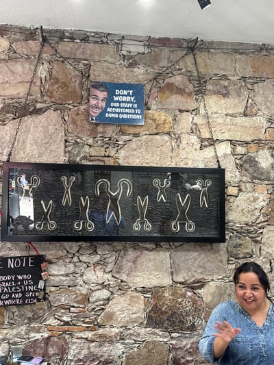 The image shows a wall in San Miguel de Allende, Mexico, constructed of roughly hewn stones. Hanging on the wall are several items: a framed collage of scissor images, a sign stating "DON'T WORRY, OUR STAFF IS ACCUSTOMED TO DUMB QUESTIONS" with a picture of a man underneath, and a small black sign titled "NOTE" with handwriting indicating someone who supports Israel and the US should go elsewhere. A woman wearing a denim shirt appears in the upper right corner of the image, smiling and partially obscuring the stonework. The items are suspended by metal chains attached to the wall. The reflection of a person in a blue shirt can be seen at the bottom of the framed collage.