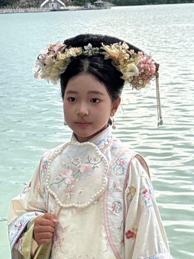 A young East Asian individual, appearing to be a child, is depicted standing outdoors against a backdrop of water and distant land structures. The individual wears a light-colored, long-sleeved garment consistent with historical Chinese attire, featuring intricate embroidery of floral patterns and small figures in shades of pink, blue, green, and red, with pink piping along the seams. A fan-shaped object with a pearl-beaded perimeter and floral embroidery is held in the right hand. The individual's dark hair is styled elaborately with a large, crescent-shaped headpiece adorned with numerous artificial flowers in hues of pink, cream, yellow, and light blue. A beaded chain with small tassels hangs from the right side of the headpiece, and pearl drop earrings are worn. The individual's expression is neutral, with eyes directed towards the viewer. The background consists of a body of light blue-green water with visible ripples. Across the water, in the distance, are trees and several architectural structures, including a prominent white building with a distinct rounded archway. This background environment identifies the location as Kunming Lake within the Summer Palace in Haidian District, China.
