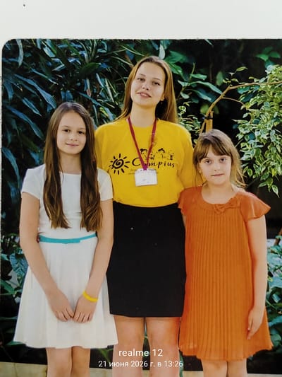 Three girls are posing for a photograph against a backdrop of green foliage. The girl on the left is wearing a white dress with a blue belt and a yellow bracelet. The middle girl is wearing a yellow t-shirt with a graphic design and a purple lanyard with an ID badge. She has her arms crossed. The girl on the right is wearing an orange dress. The photo has a timestamp in the lower right corner that reads "realme 12 21 июня 2026 г. в 13:26", which translates to "realme 12 June 21, 2026 at 13:26". The location of Shchyolkovo, Russia is not visually discernible from the image itself.