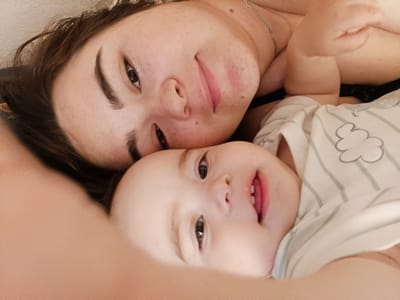 The image depicts a close-up of two individuals, likely a mother and her baby, lying down. The upper individual's face is partially visible, showing an eye, nose, and mouth. They have dark hair and a light complexion. The lower individual, a baby, is also visible, with their face facing the camera. The baby has large eyes and an open mouth, revealing tiny teeth. They are wearing a striped garment with cloud-like designs. The background is blurred, suggesting a soft focus or a shallow depth of field. The lighting appears to be natural, casting a warm glow on the subjects. There are no discernible elements indicating a specific location within the city of Посад, Russia. The scene conveys a sense of intimacy and tenderness between the two individuals.