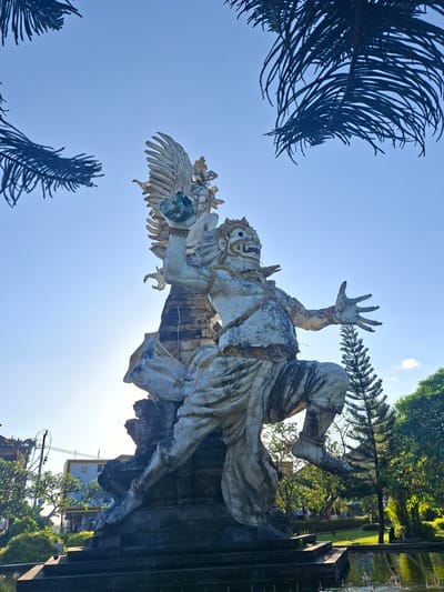 A large, weathered stone statue of a Garuda, a mythical bird-like creature from Hindu mythology, stands majestically against a clear blue sky. The statue depicts the Garuda in a dynamic pose, with one arm raised holding a globe, wings spread, and its other arm extended outwards. The creature has a fierce expression with a detailed face, adorned with a crown and jewelry. It is dressed in traditional Balinese attire, with flowing pants and a sash. The statue is situated in a park-like setting, with lush green trees and a body of water in the foreground, reflecting the sunlight. The statue is located at the Taman Festival Bali, an abandoned cultural park in Gianyar, Indonesia. The sun is bright, casting shadows and highlighting the textures of the stone.