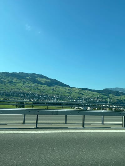 The image presents a daytime, exterior view of a landscape in Buochs, Switzerland, captured from a roadway. The foreground features a dark asphalt surface with two parallel white lines. A light grey, segmented metal guardrail extends horizontally across the lower-mid ground, supported by vertical posts that cast shadows. Beyond the guardrail, a flat, vibrant green field is visible, transitioning into a cluster of light-colored buildings with dark roofs, representing a town situated at the base of a large, green-covered hill. This hill, sparsely dotted with trees and fields, occupies the central and left background. The upper two-fifths of the frame consist of a clear, bright blue sky, with a faint, wispy white cloud in the upper-left quadrant. No individuals are visible.