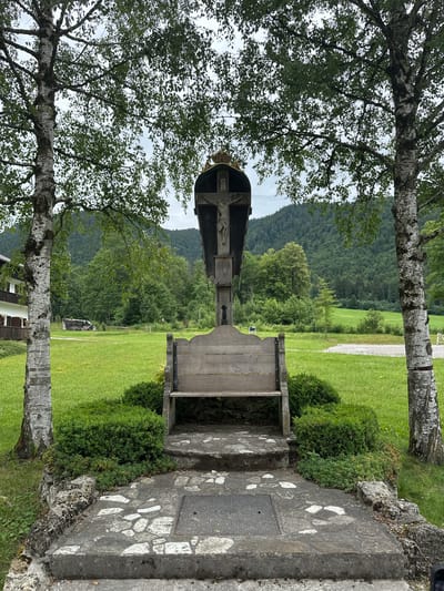 This is a full-shot, eye-level, outdoor, daytime photograph of a wayside shrine in Ettal, Germany. The shrine is situated on a paved area with stone accents and is surrounded by manicured green bushes. Behind the shrine is a wooden bench, and beyond that, a vast expanse of green grass leading to a treeline of dark green coniferous and deciduous trees, which then gives way to rolling, tree-covered hills under a cloudy sky. Two tall, slender birch trees with white bark flank the shrine. To the left, a portion of a building with a white facade and dark brown shutters is visible. The shrine itself features a wooden structure with a metal, decorative crest at the top. A depiction of Jesus Christ on the cross is visible within the shrine. The overall setting appears to be serene and rural.