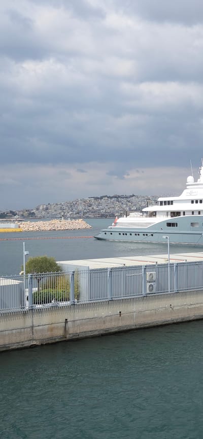 A wide-angle shot captures a marina scene in Palaio Faliro, Greece, under an overcast sky dominated by dense grey clouds. In the immediate foreground, dark blue-green water with subtle ripples is visible, bordered by a light-colored concrete quay. A grey metal fence with vertical bars and a top rail runs along the quay, behind which small green trees and light-colored, possibly industrial, structures are present. Two stacked outdoor air conditioning units are affixed to one of these structures on the right.

In the midground, a large superyacht, identified partially as "RADIANT," is moored or underway towards the right, presenting its port side. The yacht features a light grey hull and a multi-deck white superstructure, displaying a red flag on its bow and a blue and white flag (likely Greek) on its stern. An orange floating line or boom extends from the yacht's bow area towards the left. To the yacht's left, a light-colored rock breakwater is present, topped by a small white and black navigational beacon or lighthouse. Behind the breakwater, a section of a large yellow marine vessel or barge is partially visible.

The background reveals a densely populated urban hillside, characteristic of the city, covered with numerous white and light-colored buildings extending towards the horizon under the heavy cloud cover. No individuals are discernible in the frame. The scene is static, depicting a calm harbor environment.