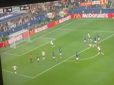 The image shows a football match on a green grass field, with players in white and blue uniforms. There are goalposts and a net visible. Spectators are in the background, seated in stands. A red advertising board with the McDonald's logo is present along the side of the field. In the upper left corner, a scoreboard displays "FRA 0 0 ESP". A banner in the background reads "НА ЖИВО" and "FC". The scene appears to be a professional football game, likely in a stadium. The specific location in Sredets, Bulgaria cannot be determined from the provided image alone, as it lacks any identifying landmarks or text specific to that city.