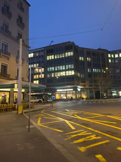 The image captures an urban street scene at dusk or night in Genève, Switzerland. A deep blue sky hangs above a cityscape dominated by buildings and artificial light. On the right, a large modern building, likely an office block, stands tall with many windows brightly illuminated, reflecting light. At its ground level, a "STARBUCKS COFFEE" shop is prominently visible, glowing with interior lights, and a few individuals can be seen inside.
In front of the buildings, a wide paved road is marked with clear yellow lines, including a section with cross-hatching and the word "TAXI" painted on the asphalt, indicating a designated taxi stand. A public transport vehicle, possibly a bus or tram, is partially visible on the street, with the letters "IVE" discernible on its side.
To the left, an older, possibly residential, building with architectural details is partially captured. Overhead utility wires are visible against the dark sky. Below the older building, stacked tables and chairs suggest an outdoor seating area for another establishment. The overall atmosphere is calm and quiet, characteristic of an urban evening.