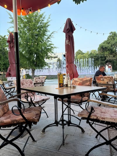 The image depicts an outdoor seating area of a cafe, likely in Svishtov, Bulgaria, given the context. In the foreground, a square wooden table with a dark metal base is set with salt and pepper shakers, a sugar dispenser, and a small menu. Surrounding the table are chairs with ornate black metal frames and cushions patterned with floral and abstract designs. One chair in the foreground is angled towards the viewer, revealing its decorative cushion. In the middle ground, a man with short dark hair is seated at a table, facing to the right and looking away from the camera. He is wearing a dark, patterned t-shirt and appears to be a patron. Behind the seating area, a fountain with multiple spouts of water is visible, illuminated by white and purple lights. Trees with lush green foliage line the background, and beyond them, buildings can be seen. String lights with Edison bulbs are hung across the top of the scene, adding a decorative element to the ambiance. Two large, burgundy patio umbrellas are partially visible, one folded and the other angled. The ground is paved with stone tiles. The overall impression is a relaxed outdoor dining setting, possibly in a public square, with the fountain as a prominent feature.