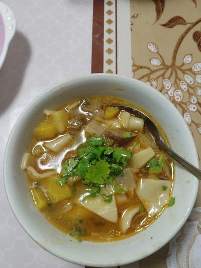 A close-up shot shows a bowl of soup filled with noodles, potatoes, and meat. The soup is in a white bowl with a silver spoon resting on the edge. The background shows a tablecloth with a floral pattern and a table runner. The lighting is warm and natural, suggesting it might be daytime. The food appears to be a hearty meal, possibly a traditional Kyrgyz dish given the location context of Kaarman, Kyrgyzstan. There is no visible text in the image.