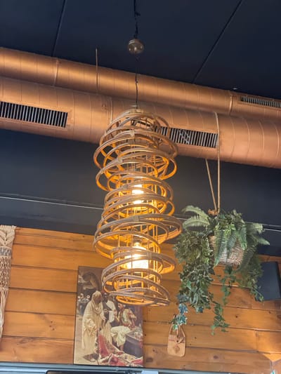 This media file captures an indoor scene, likely a restaurant or commercial space, characterized by its industrial-chic and bohemian decor. Prominently featured are two large, copper-colored ventilation ducts, positioned vertically and running parallel to each other. These ducts have horizontal ventilation grilles and are a dominant element of the industrial aesthetic.
Hanging from the ceiling is a distinctive chandelier made of woven natural material, possibly rattan or bamboo, forming a cylindrical shape with multiple concentric rings. Inside the chandelier, several light bulbs are illuminated, casting a warm glow. This bohemian element contrasts with the industrial ducts.
Above and slightly to the right of the chandelier, a hanging planter with lush green artificial plants, including ferns and ivy, adds a touch of biophilic design. These plants are housed in a woven basket, further enhancing the bohemian theme.
On the wooden-paneled wall to the right of the image, a framed artwork is visible. It depicts a religious or historical scene with figures dressed in flowing robes, rendered in muted tones. The artwork adds a classical or vintage touch to the space. Below this artwork, a section of a woven textile, possibly a rug or wall hanging, is visible.
A small, light-colored object, possibly a decorative item or a small bag, hangs from the wall near the plants.
The lighting suggests it could be daytime, but the interior setting makes it difficult to determine the time of day definitively. There are no clear indicators of weather or specific time, but the overall ambiance is warm and inviting. The location context provided, Sant Adrià de Besòs, Spain, does not offer any visually discernible clues within the image itself. No visible text is present in the image.