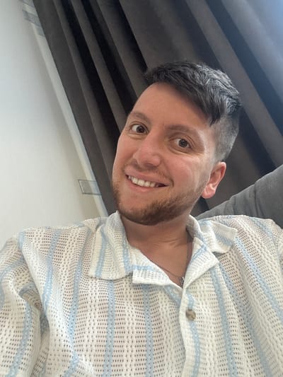 The image shows a close-up of a man's face and upper torso. He has brown eyes, dark hair, and a beard. He is smiling and wearing a white, short-sleeved shirt with blue stripes. The background is blurred and appears to be a room with dark curtains on the left and a grey upholstered chair on the right. There is a white wall to the left of the man. The location is identified as Мирный, Russia.