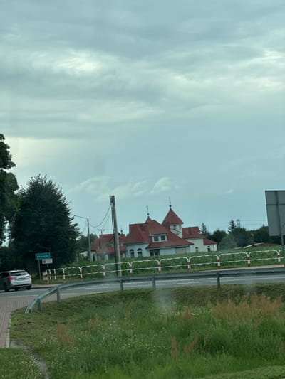 The image shows a cloudy sky over a rural landscape in Terespotockie, Poland. A white car is parked on the side of the road, next to a green sign that reads "Urbanowo." In the background, a large, white house with a red roof and multiple turrets is visible. A metal guardrail separates the road from a grassy field with wildflowers. Trees are scattered throughout the scene. The overall impression is of a quiet, suburban or rural area.