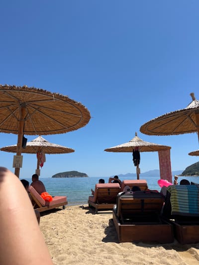 The image depicts a sunny day at a beach in Νέα Ηρακλείτσα, Greece. Several thatched umbrellas provide shade over lounge chairs on a sandy shore. The calm, blue sea stretches to the horizon, with a small, tree-covered island visible in the distance. Mountains are silhouetted against the clear blue sky in the background. Several people are relaxing on the lounge chairs, some partially visible in the foreground, while others are seated further back. Beach towels and inflatable rings are visible on some of the chairs. The atmosphere appears relaxed and leisurely.