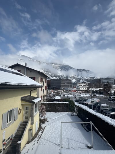 The image depicts a snow-covered winter scene in Visp, Switzerland, under a partially cloudy blue sky. In the immediate foreground on the left, a light yellow multi-story residential building features a snow-laden gabled roof, grey shutters on white-framed windows, and a front entrance accessed by stone steps, adorned with a wreath on the white door. Fresh footprints are visible in the snow along the side of this building. To its right, a snow-covered yard contains a white metal frame structure and a dark green hedge delineating the property. Further back, another light-colored residential building with red shutters and a snow-covered roof is visible. The midground shows a paved area serving as a parking lot, with multiple vehicles, including sedans, SUVs, and a white commercial van, partially covered in snow. Beyond the parking area, several urban structures are discernible, including a prominent dark grey, multi-story modern building with horizontal elements and a traditional white building with a dark roof. The background is dominated by towering, snow-covered mountains, partially obscured by clouds or mist. No individuals are present in the scene.