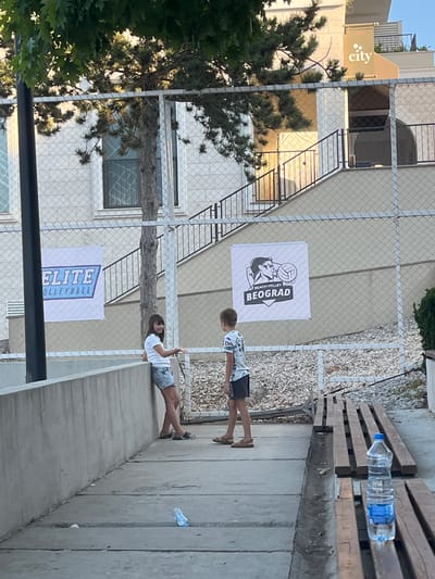This image captures a sunny outdoor scene, likely in Gošići, Montenegro, featuring a beach volleyball court area. In the foreground, a concrete pathway runs alongside a low concrete wall. A young girl in a white t-shirt and denim shorts leans against the wall, looking towards the camera and smiling. To her right, a young boy wearing a patterned t-shirt and shorts stands facing away, also looking towards the court. Behind them, a chain-link fence encloses the area.
Two large banners are visible attached to the fence. The banner on the left reads "ELITE VOLLEYBALL" in blue lettering. The banner on the right features a graphic with a volleyball player and the text "BEACH VOLLEY BEOGRAD". The background shows a light-colored building with arched windows and staircases leading up to it. A large tree with sparse foliage is positioned in the middle ground, partially obscuring the view of the building.
In the bottom right corner, wooden benches are arranged, with a plastic bottle of water placed on one of them. The ground is covered with small white stones. The overall environment suggests a recreational area, possibly part of a sports complex or a public space near the coast.