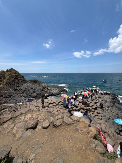 The image depicts a rocky coastal landscape with a clear blue sky and scattered white clouds. The foreground is dominated by a dark, hexagonal rock formation that extends into the ocean. A group of people are gathered on the rocks, many holding colorful umbrellas for shade. Some individuals are standing and observing the scenery, while others are seated. The sea is a deep blue, with a few small boats visible in the distance. The overall environment suggests a popular tourist destination, possibly within Phú Lương, Vietnam, known for its unique geological features.