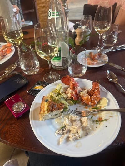 This image depicts an indoor dining scene at a restaurant or similar establishment in Cernay-la-Ville, France. A dark wooden table, likely round or oval, is laden with the aftermath of a meal.
In the foreground, a large white ceramic plate contains partially consumed food: a wedge of quiche, two large lobster tails with some shell remnants, a small portion of lettuce salad, and a pile of shredded white meat in a creamy sauce. A silver fork rests on the plate. To the left of this plate, a black smartphone displaying the time "3:13" on its screen is positioned face down. Adjacent to the smartphone is a magenta Sony Cyber-shot digital camera, model "Claremont." A small black sachet, possibly for sauce or cleaning, is also visible.
Scattered across the table are several clear stemmed wine glasses, most containing varying levels of white wine, and several clear water glasses, some full and some empty. A clear glass bottle of Badoit sparkling water, largely empty, stands near the center. Further back on the table, a smaller white ceramic plate holds remnants of several eaten shrimp. Silverware, including knives and spoons, is also present on the table.
Decorative elements include a few red berries and green leaves scattered on the table surface. A light-colored wooden salt or pepper grinder is visible in the background. The background is blurred, showing soft pink upholstered seating and a wicker object. Partially visible human figures are present, one appearing to be wearing a white uniform or apron, suggesting staff or other diners. The overall scene indicates a recently concluded or ongoing multi-course meal.