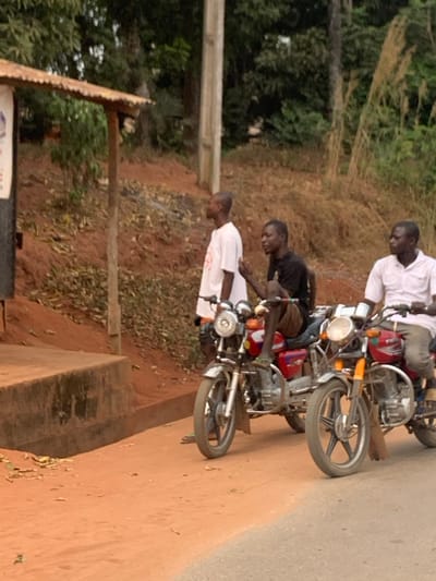 The scene in Aguleri, Nigeria, depicts three adult males and two motorcycles on a reddish-brown dirt path alongside a grey paved road. On the far left, a male subject wearing a white t-shirt and light-colored shorts stands facing right, positioned next to a concrete curb and a dilapidated structure with a corrugated metal roof. To his right, a second male subject, dressed in a black t-shirt and brown shorts, sits astride a red motorcycle, facing left with his left hand raised in a gestural posture. A third male subject, wearing a white collared shirt and light-colored trousers, is seated on a second red motorcycle to the right of the first, facing slightly left. Both motorcycles feature chrome components and visible engines. The immediate background consists of a sloping bank of reddish-brown earth covered with dry, sparse vegetation, topped by dense green trees. A wooden utility pole is visible in the background, left of center. The lighting suggests daytime.