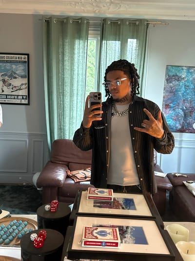 A person with dreadlocks and glasses is taking a selfie in a room with neutral-colored walls and curtains. The person is wearing a white t-shirt, a black pinstripe jacket, and a silver chain. They are holding a smartphone in their left hand and making a hand gesture. In the background, there's a painting on the wall, a leather chair, framed pictures with Bicycle brand playing cards attached to them, dice, and a decorative bowl with blue marbles. There is no indication of location.