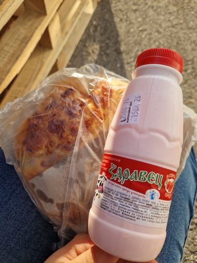 A person holds a bottle of pink strawberry yogurt and a wrapped pastry in front of a wooden pallet structure. The yogurt bottle is white with a red cap and a label that reads "Здравец" in green lettering. The label also includes images of strawberries and a cow. A sticker on the bottle has Cyrillic text and dates. The pastry appears to be a savory pie with a golden-brown crust, wrapped in clear plastic. The scene is outdoors, with sunlight casting shadows. The image context suggests this may be related to a visit to Bahovitsa, Bulgaria, given the mention of the location in the prompt.