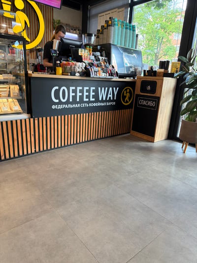 This media file shows a counter at a "COFFEE WAY" cafe in Tikhoretsk, Russia. A female barista is visible behind the counter, interacting with a computer screen and serving customers. The cafe appears to be well-lit, with natural light coming in from a large window overlooking trees and buildings. The counter area features a display of coffee cups, various packaged snacks, and a large "COFFEE WAY" sign with Russian text that translates to "FEDERAL CHAIN OF COFFEE SHOPS". There is also a "THANK YOU" sign in Russian on a wooden structure above the counter. The flooring is tiled, and the overall atmosphere suggests a busy but pleasant coffee shop setting. The time of day appears to be daytime, based on the natural light.